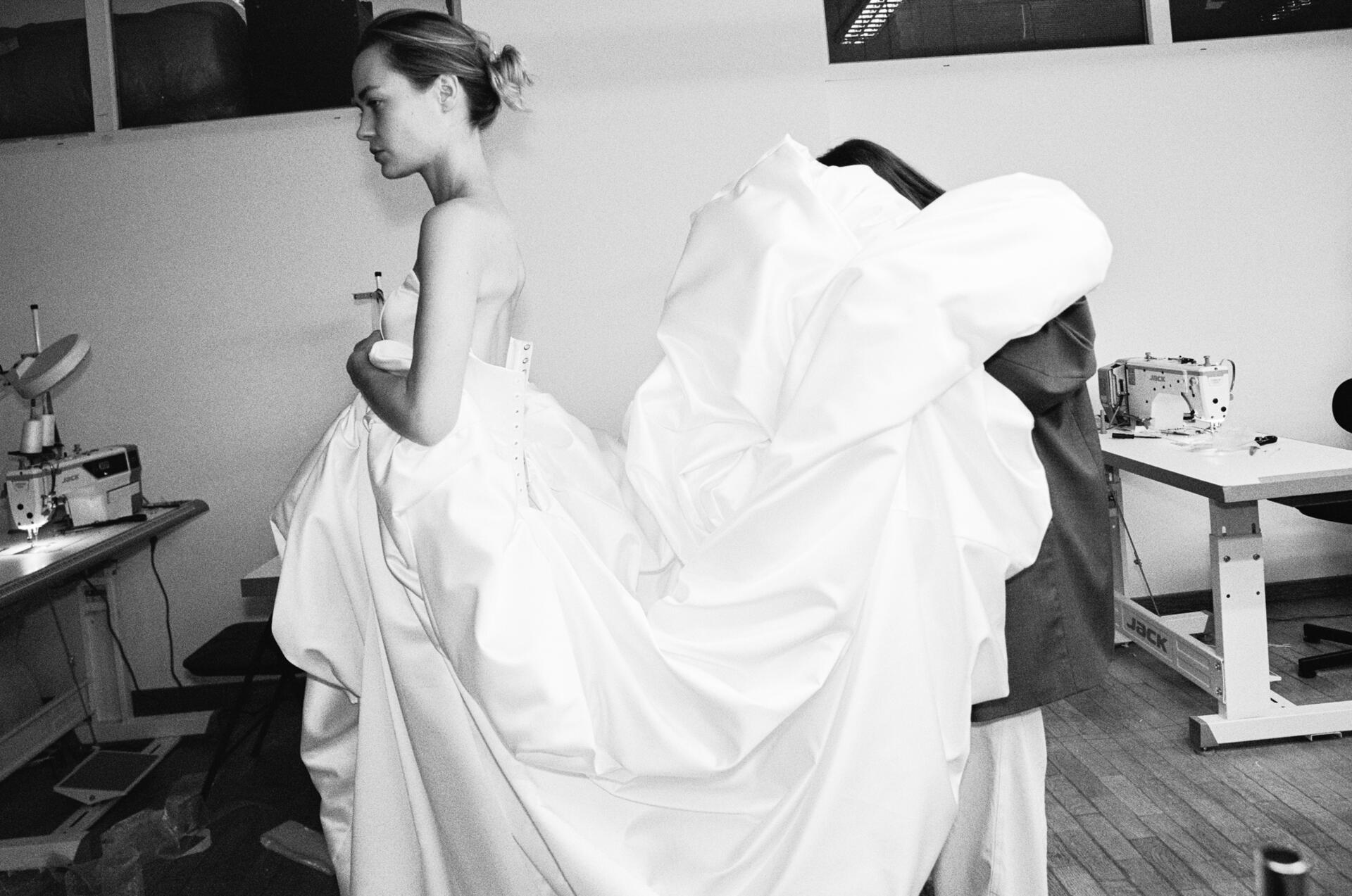 A bride in a strapless gown stands in the sewing atelier while a seamstress gathers up her train