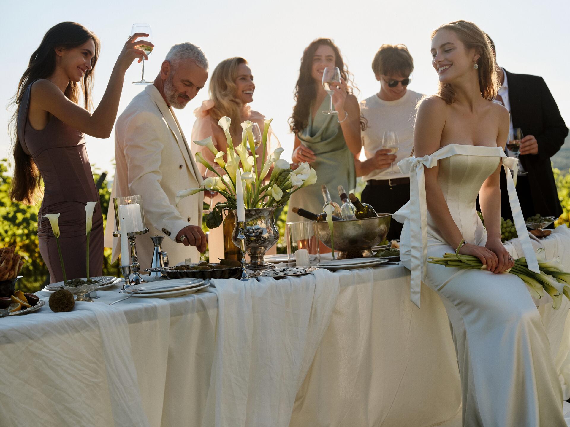 A bride in an off-the-shoulder satin gown sits on a long table set with calla lilies while friends raise their glasses in the sun