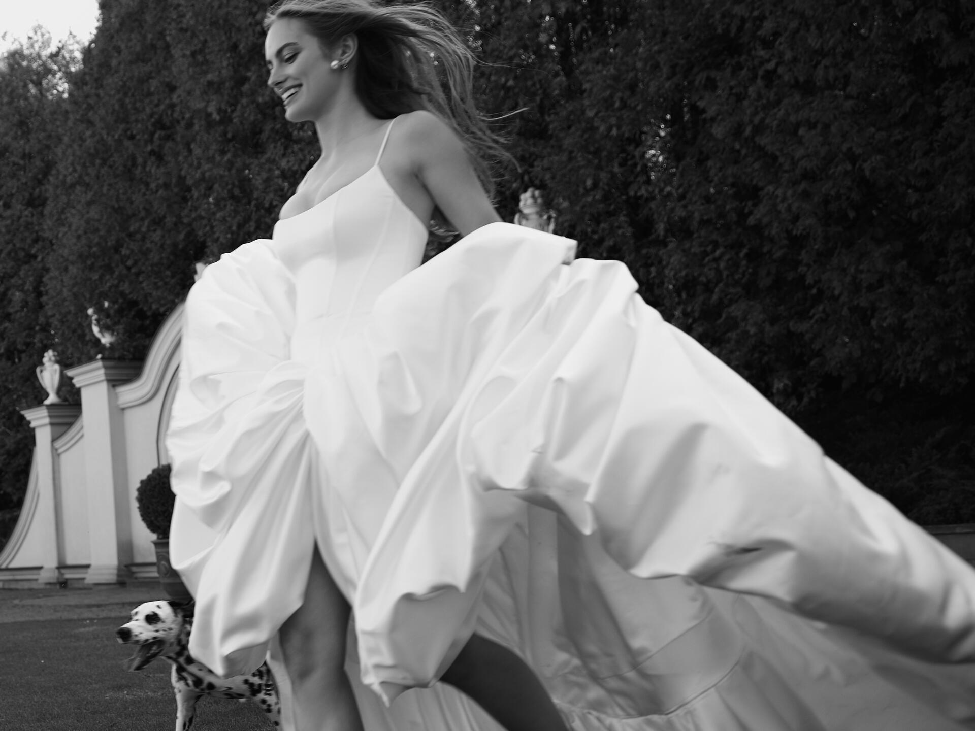 A smiling bride in a full white gown twirls in front of a garden hedge